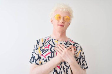Renkli tişört ve güneş gözlüğü takan genç albino adam izole edilmiş kırmızı arkaplanda elleri göğsünde, gözleri kapalı ve yüzünde minnet dolu bir jest ile gülümsüyor. Sağlık kavramı.