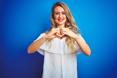 Mavi izole edilmiş arka planda duran beyaz elbiseli genç ve güzel bir kadın. Aşkla gülümsüyor. Elleriyle kalp sembolü ve şekli gösteriyor. Romantik konsept.