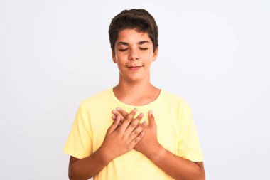 Yakışıklı genç çocuk izole edilmiş beyaz arka planda duruyor elleri göğsünde, gözleri kapalı ve yüzünde minnet dolu bir jest ile gülümsüyor. Sağlık kavramı.