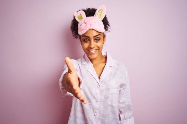 Genç afro-amerikan kadın pijama giyip maske takıyor pembe arka planda dostça gülümseyerek selamlama ve hoş geldin diyor. Başarılı bir iş..