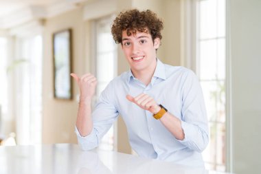 Kıvırcık saçlı genç bir iş adamı. Arkasını işaret ediyor. El ve baş parmakları havada, kendine güvenen.