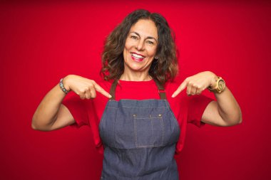 Kırmızı izole edilmiş arka planda önlük giyen orta yaşlı bir kadın yüzünde bir gülümsemeyle gururla ve mutlu bir şekilde kendini işaret ediyor..