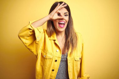 Sarı arka plan üzerinde duran kot ceket giyen genç ve güzel bir kadın el hareketleriyle gülümsüyor ve mutlu yüzüyle parmaklarının arasından bakıyor..
