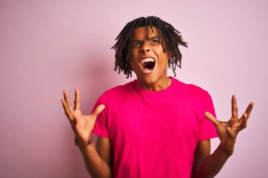 Rastalı tişörtlü Afro-Amerikan bir adam pembe arka planda dikiliyor deli gibi bağırıyor ve agresif bir ifadeyle bağırıyor ve kollarını kaldırıyor. Hayal kırıklığı konsepti.