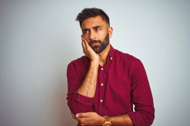 Kırmızı şık gömlek giyen genç Hintli adam gri arka planda duruyor. Kollarını kavuşturup depresyondan sıkılmış ve yorgun görünüyor..