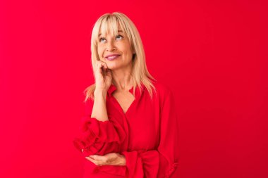 Orta yaşlı bir kadın zarif bir gömlek giyer, izole edilmiş kırmızı arka plan üzerinde durur, elleri çenesinde soru sorarken, düşünceli bir ifade takınır. Düşünceli bir yüzle gülümsüyor. Şüphe kavramı.