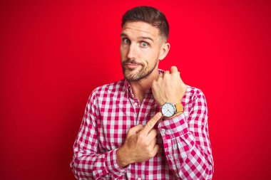Kırmızı izole edilmiş arka plan üzerinde genç yakışıklı bir adam. Aceleyle saate, sabırsızlığa ve rahat bir ifadeyle kameraya bakıyor.