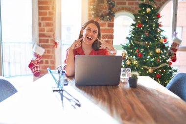 Güzel bir kadın evde dizüstü bilgisayarla oturmuş Noel ağacının etrafında gülüyor ve parmaklarını ve ağzını göstererek gülümsüyor. Diş sağlığı konsepti.