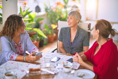 Orta yaşlı kadınlarla buluşup, öğle yemeği yiyip, kahve içiyorlar. Olgun arkadaşlar mutlu bir şekilde gülümsüyor. Güneşli bir günde evde akıllı telefon kullanıyorlar.