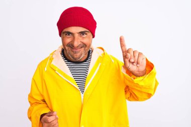Yağmurluk ve yün şapka giyen orta yaşlı bir adam izole edilmiş beyaz arka plan üzerinde duruyor ve bir numaralı parmağını gösterirken kendinden emin ve mutlu bir şekilde gülümsüyor..