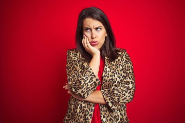 Genç, güzel, esmer, leopar desenli bir ceket giyen, kırmızı izole bir arka plan giyen kadın kollarını kavuşturarak depresyondan sıkılmış ve yorgun gözüktüğünü düşünüyor..