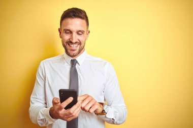 Genç yakışıklı iş adamı, sarı izole edilmiş arka planda akıllı telefon kullanıyor. Mutlu bir yüzü var. Kendine güvenen bir gülümsemeyle dişlerini gösteriyor.