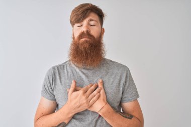 Genç kızıl İrlandalı adam kapalı gözleri ve yüzünde minnettar jest ile göğüs üzerinde gülümseyerek izole gri arka plan üzerinde duran t-shirt giyiyor. Sağlık kavramı.