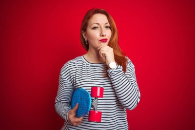 Kızıl saçlı genç bir kız izole edilmiş kırmızı arka planda paten kayıyor. Ciddi bir yüz ifadesi var. Soru sormayı düşünüyor.