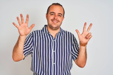 Mavi çizgili tişört giyen genç adam izole edilmiş beyaz arka plan üzerinde duruyor ve 8 numaralı parmaklarıyla işaret ederken kendinden emin ve mutlu bir şekilde gülümsüyor..