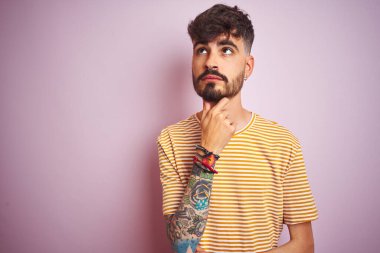 Sarı çizgili tişört giyen genç bir adam soyutlanmış pembe arka planda duruyor ve elleri çenesinde soru sormayı düşünüyor, düşünceli bir ifade. Düşünceli bir yüzle gülümsüyor. Şüphe kavramı.