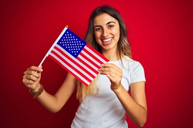 Amerika Birleşik Devletleri bayrağını kırmızı izole edilmiş arka planda tutan genç bir kadın mutlu bir yüzle ayakta duruyor ve gülümsüyor.
