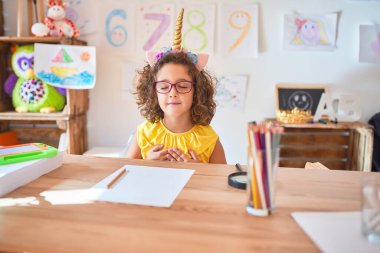 Gözlüklü ve tek boynuzlu at diademi takmış güzel bir çocuk anaokulunda masasında oturmuş elleri göğsünde, gözleri kapalı ve yüzünde minnet dolu bir jest ile gülümsüyor. Sağlık kavramı.