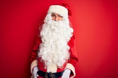 Noel Baba kostümü giymiş orta yaşlı yakışıklı bir adam izole edilmiş kırmızı arka planda dikiliyor komik bir ifadeyle dilini dışarı çıkarıyor. Duygu kavramı.