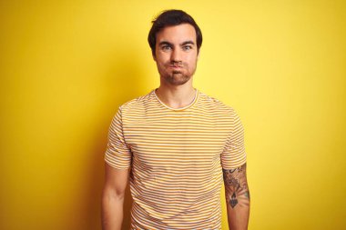 Dövmeli, çizgili tişörtlü, izole edilmiş sarı arka planda, komik yüzlü, şişkin yanaklı genç bir adam. Havayla şişmiş ağız, çılgın bir ifade..