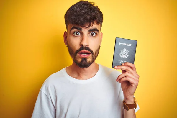 Young man with tattoo wearing United States USA passport over isolated yellow background scared in shock with a surprise face, afraid and excited with fear expression