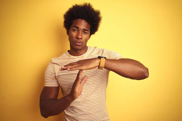 American man with afro hair wearing striped t-shirt standing over isolated yellow background Doing time out gesture with hands, frustrated and serious face