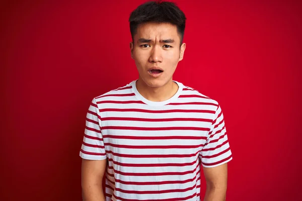 若いアジア系中国人男性の縞模様のTシャツの上に立って孤立した赤の背景を恐れていると驚きの表現でショックを受けました 恐怖と興奮した顔 — ストック写真