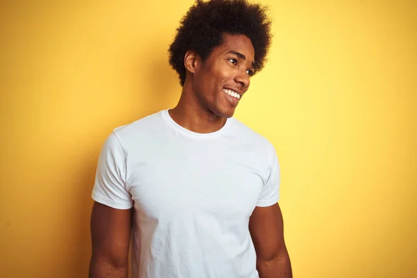 American man with afro hair wearing white t-shirt standing over isolated yellow background looking away to side with smile on face, natural expression. Laughing confident.