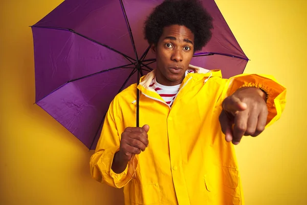 Afro american man wearing rain coat and umbrella standing over isolated yellow background pointing with finger to the camera and to you, hand sign, positive and confident gesture from the front
