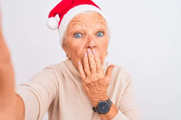 Grey-haired woman wearing Crhistmas Santa hat make selfie over isolated white background cover mouth with hand shocked with shame for mistake, expression of fear, scared in silence, secret concept