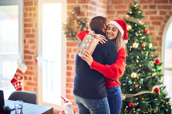 Young beautiful couple smiling happy and confident. Standing holding gift and hugging around christmas tree at home