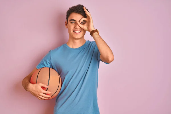 Teenager boy holding professional basket ball over isolated pink background with happy face smiling doing ok sign with hand on eye looking through fingers