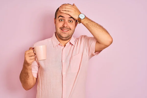 Young man drinking cup of coffee standing over isolated pink background stressed with hand on head, shocked with shame and surprise face, angry and frustrated. Fear and upset for mistake.