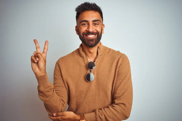 Indian man wearing elegant sweater and sunglasses standing over isolated white background smiling with happy face winking at the camera doing victory sign. Number two.