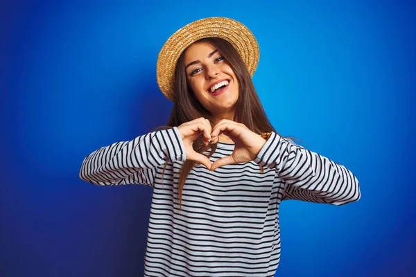 Young beautiful woman wearing navy striped t-shirt and hat over isolated blue background smiling in love showing heart symbol and shape with hands. Romantic concept.