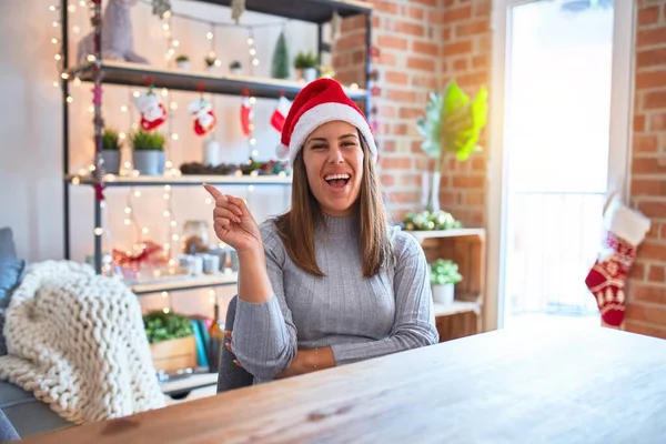 カメラを見ている側に手と指で指して 顔に大きな笑顔で家のテーブルに座ってクリスマスの帽子をかぶって若い美しい女性 — ストック写真