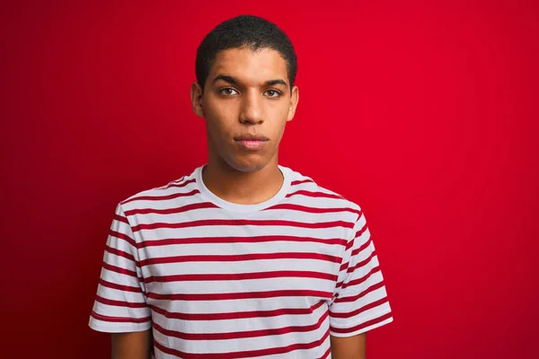 孤立した赤い背景にストライプのTシャツを着た若いハンサムなアラブ人男性が顔に深刻な表情でリラックスしました シンプルで自然なカメラを見て — ストック写真