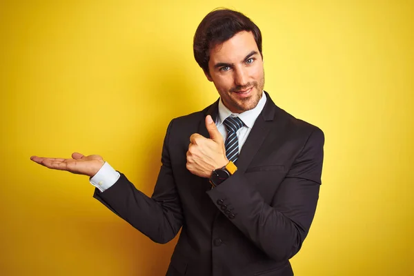 若いですハンサムなビジネスマンは スーツを着て 孤立した黄色の背景の上に立ってタイ手のひらの手を示し 親指でOkジェスチャーを行います 笑顔と陽気な — ストック写真