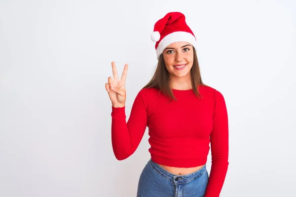 若いです美しいです女の子身に着けていますクリスマスサンタ帽子立って上の隔離された白い背景ショーと指差す指数2ながら笑顔自信と幸せ — ストック写真