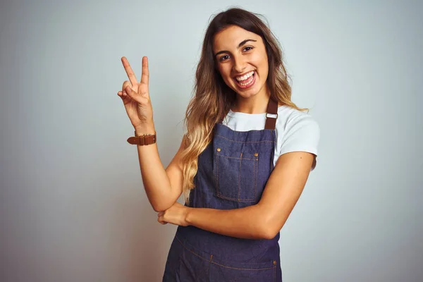 Young beautiful woman wearing apron over grey isolated background smiling with happy face winking at the camera doing victory sign. Number two.