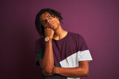 Rastgele tişörtler giyen Afro bir adam izole edilmiş mor arka plan üzerinde duruyor ve elleri çenesinde soru sormayı düşünüyor, düşünceli bir yüz ifadesi var. Düşünceli bir yüzle gülümsüyor. Şüphe kavramı.
