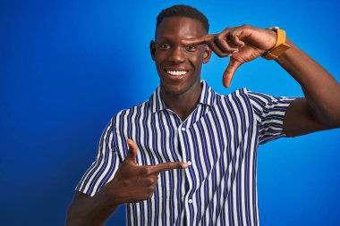 İzole edilmiş mavi arka plan üzerinde duran çizgili bir tişört giyen Afrikalı Amerikalı adam gülümsüyor ve elleri ve parmakları mutlu yüzlü bir çerçeve yapıyor. Yaratıcılık ve fotoğrafçılık kavramı.
