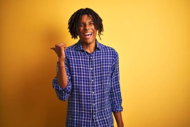 Rastgele tişörtlü Afro adam izole edilmiş sarı arka planda dikiliyor mutlu yüzüyle gülümsüyor ve başparmağıyla yan tarafı işaret ediyor..