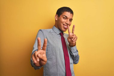 Yakışıklı Arap iş adamı gömleği ve kravat takıp izole edilmiş sarı arka planda gülümsüyor ve parmaklarını zafer işareti yaparken gösteren kameraya bakıyor. İki numara..