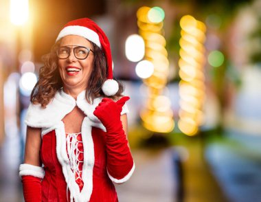 Orta yaşlı kadın, Noel Baba kostümü giyiyor, gülümsüyor, gülümsüyor ve başparmağıyla yan tarafa bakıyor..