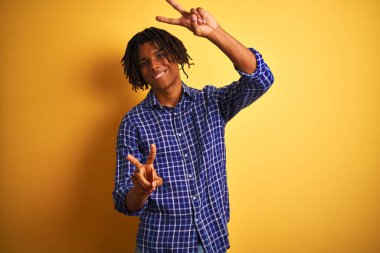 Rastgele tişörtlü Afro adam izole edilmiş sarı arka planda dikilip kameraya bakıp zafer işareti yaparken gülümsüyor. İki numara..