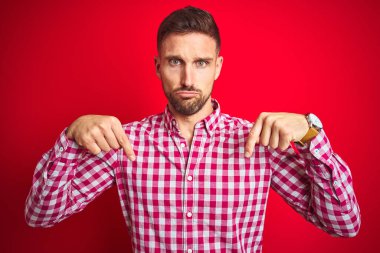 Kırmızı izole edilmiş arka plan üzerinde genç yakışıklı adam üzgün ve üzgün görünüyor, parmaklarıyla yönü gösteriyor, mutsuz ve depresif..