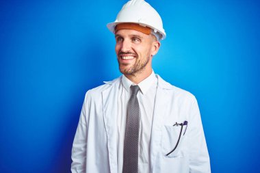 Güvenlik kaskı takan genç ve yakışıklı bir mühendis mavi izole edilmiş bir arka planda yüzünde gülümseme, doğal bir ifade ile yan yana bakıyor. Kendinden emin gülen.
