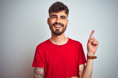 İzole edilmiş beyaz arka plan üzerinde duran kırmızı tişörtlü genç adam yüzünde kocaman bir gülümseme, eli ve parmağı kameraya bakıyor..