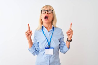 Gözlüklü ve kimlik kartı takan orta yaşlı bir iş kadını beyaz arka planda şaşkın şaşkın ve şaşırmış bir şekilde parmaklarıyla işaret ediyor ve kollarını kaldırıyordu..